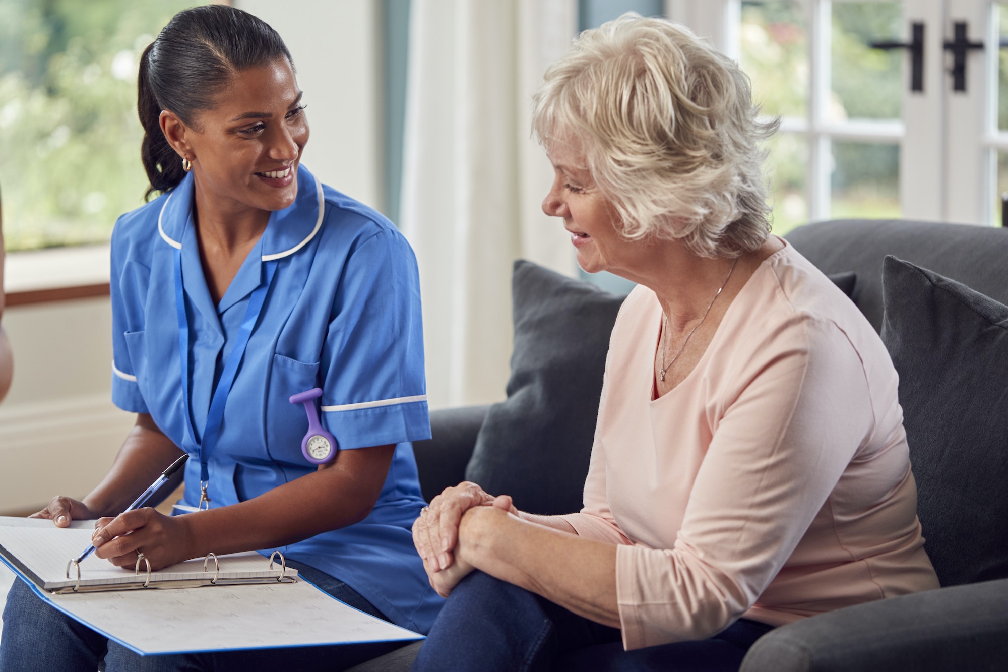 Senior Woman At Home Talking To Female Nurse Or Care Worker In Uniform Making Notes In Folder